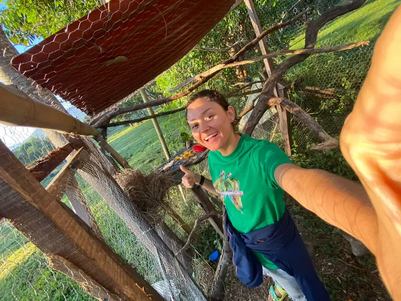 Una joven con playera verde sonríe y se toma una selfi dentro de un aviario al aire libre, entre ramas, malla de alambre y una charola con fruta.