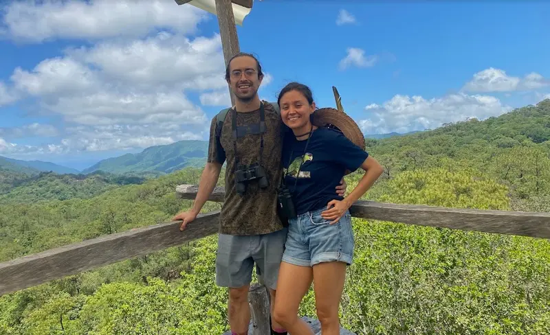 Un hombre y una mujer con binoculares sonríen en un mirador de madera frente a cerros cubiertos de bosque, bajo un cielo azul con nubes.