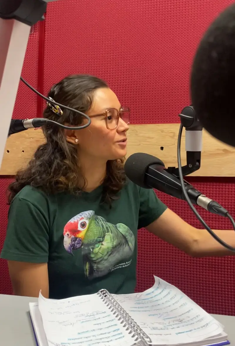 Una mujer con lentes habla ante un micrófono en una cabina de radio, con una playera verde con un loro estampado y un cuaderno abierto frente a ella.